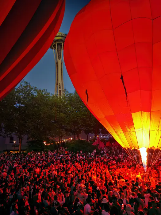 A crowd watches a red hot air balloon fire its burner beneath the Space Needle
