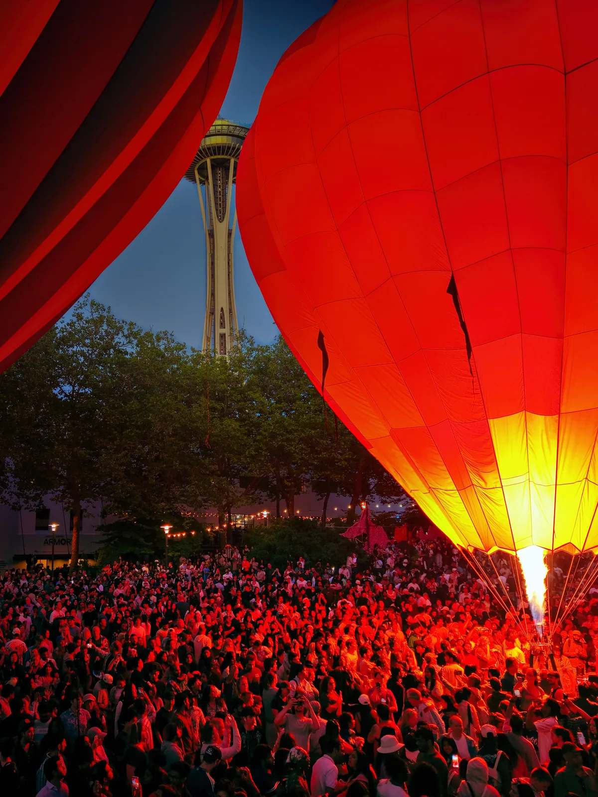 A crowd watches a red hot air balloon fire its burner beneath the Space Needle